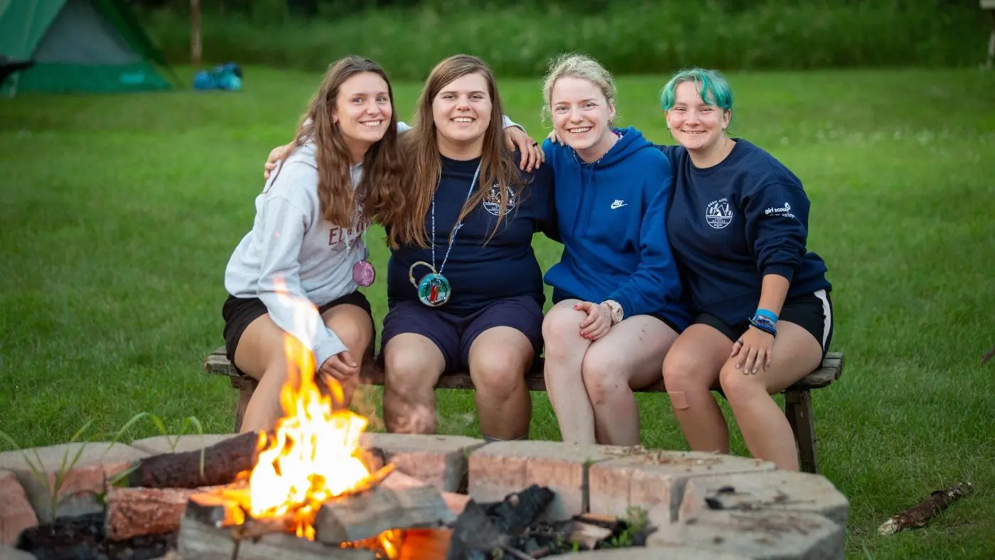 Four female friends sitting on a bench around a camp fire, all smiling with their arms around each other.