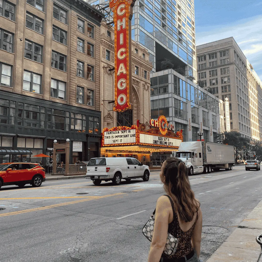 Girl standing on the side of the street looking up at a neon 'Chicago' sign on the side of a building.