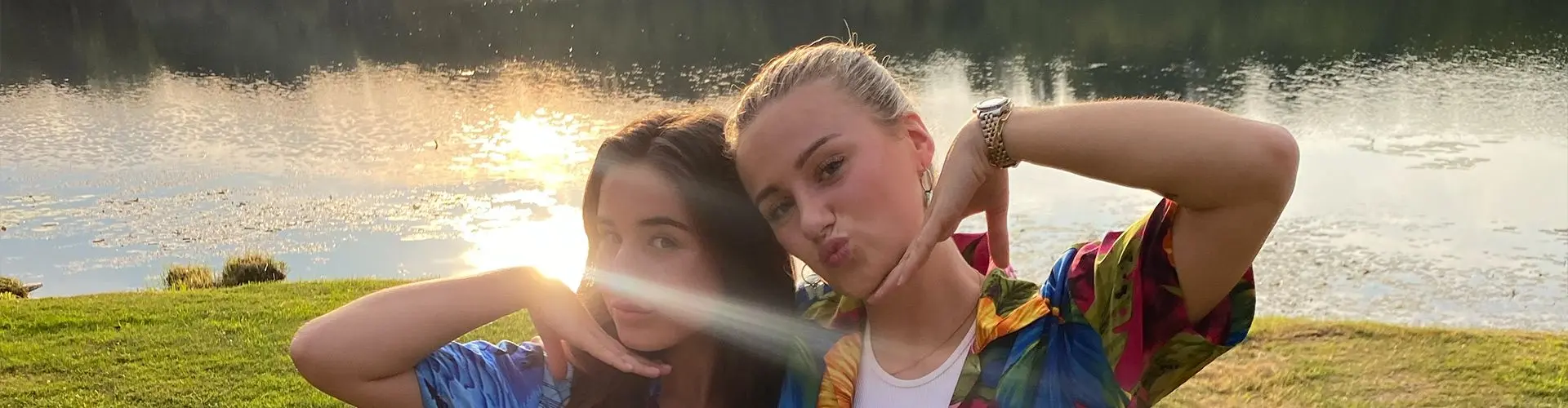 2 girls pose with hand below chin on sunny evening with grass and a lake in the background.