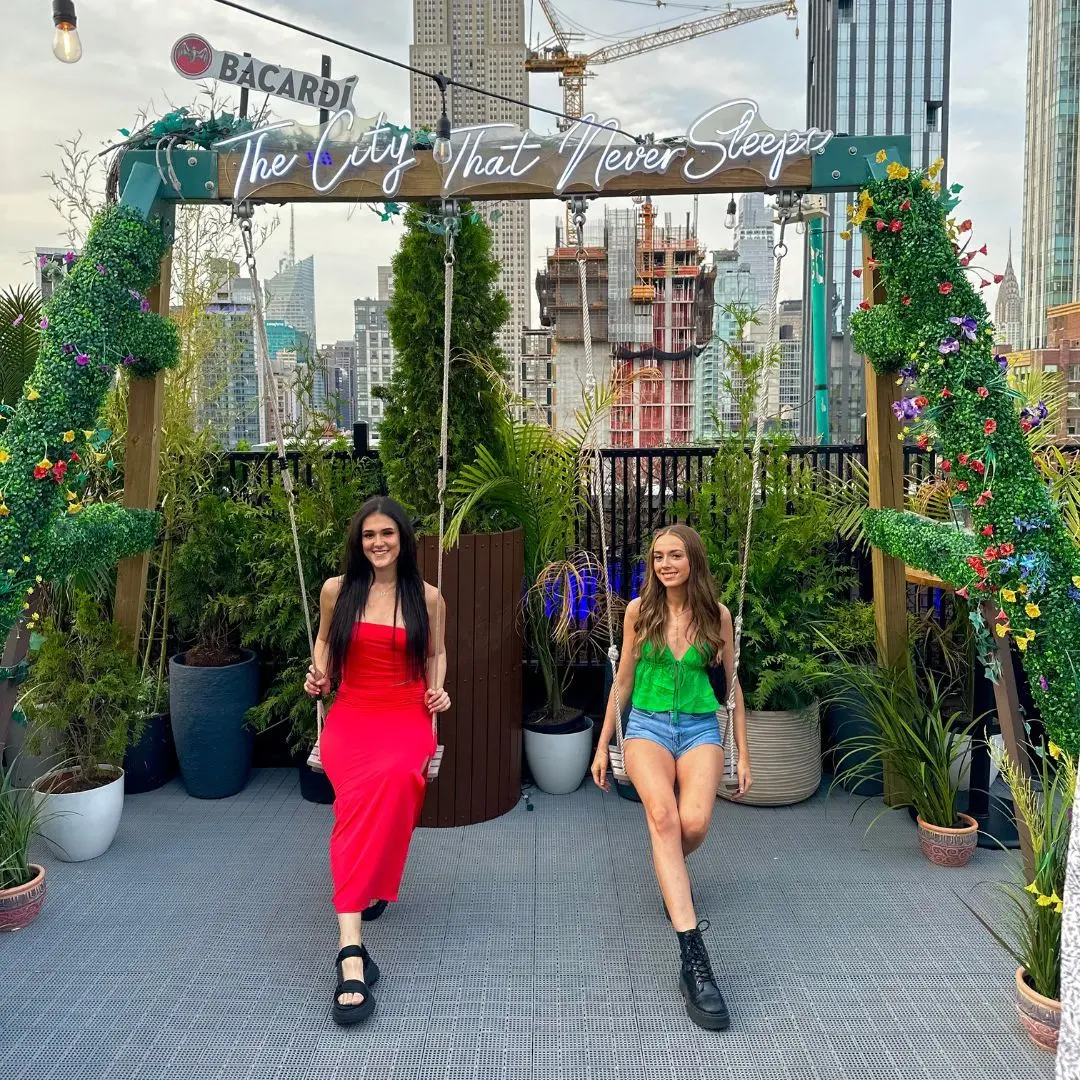 Two girls in evening outfits sitting on a swing on a rooftop in New York. The swing frame is covered in flowers and branches, and features a neon sign at the top that says 'The City That Never Sleeps'